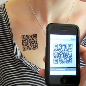 May include: A person with a black and white QR code tattoo on their chest, holding a black smartphone with the same QR code displayed on the screen. The phone screen also shows the URL "http://www.barcodeart.com".