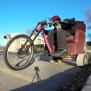 May include: A person riding a pink and black tricycle with a large, red armchair as the seat. The tricycle has a large front wheel and two smaller rear wheels.