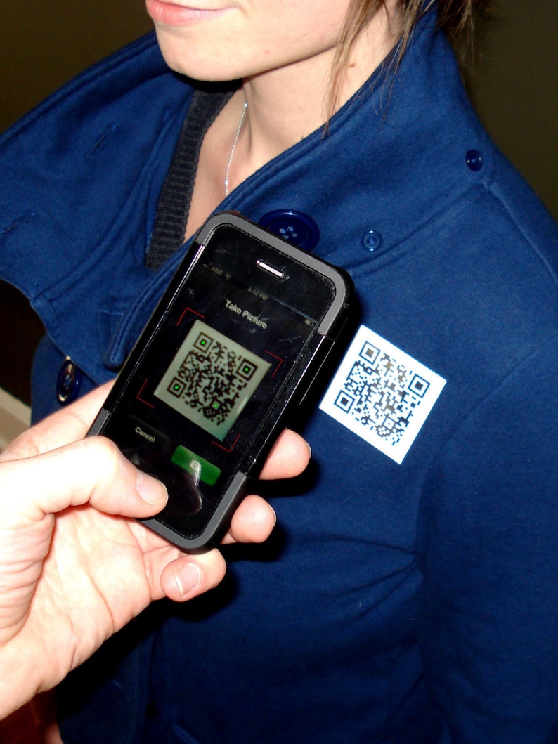 Puede incluir: Un tel&eacute;fono inteligente negro con una funda azul y un c&oacute;digo QR que se muestra en la pantalla. El texto "Take Picture" es visible en la pantalla. El tel&eacute;fono est&aacute; siendo sostenido por una persona que lleva una chaqueta azul.