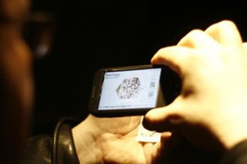 Puede incluir: Una persona usando un tel&eacute;fono inteligente para escanear un c&oacute;digo de barras en una tarjeta blanca. El c&oacute;digo de barras es blanco y negro y tiene el texto "WWW.FANDANGO.COM" impreso debajo.