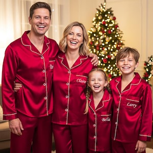 Puede incluir: Una familia de cuatro personas con pijamas de satén rojo a juego con ribete blanco, posando frente a un árbol de Navidad. Los pijamas tienen nombres bordados en la parte delantera. El fondo incluye una chimenea y decoraciones navideñas.