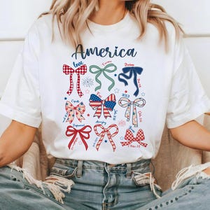 May include: White t-shirt featuring the word "America" in blue script above a collection of colorful bow illustrations. The bows are in red, white, and blue, with various patterns and text elements. The shirt is worn by a person.