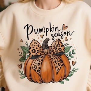 Puede incluir: Sudadera color crema con una calabaza naranja y un lazo con estampado de leopardo. Las palabras "Pumpkin season" están escritas encima de la calabaza en escritura negra, con corazones y hojas. Un diseño otoñal.