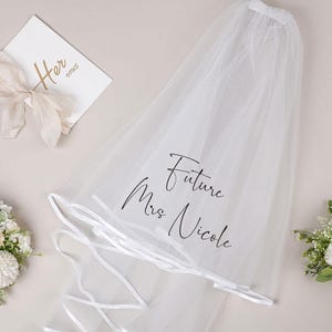 Pode incluir: Véu de noiva branco com acabamento em cetim e o texto "Future Mrs. Nicole" escrito em caligrafia preta.