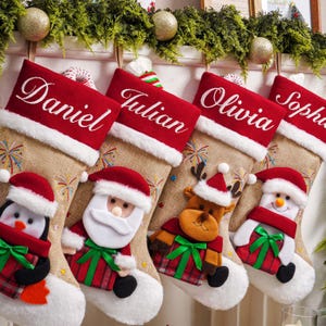 Könnte beinhalten: Vier Weihnachtsstrümpfe aus Jute mit roten Bündchen, bestickt mit den Namen Daniel, Julian, Olivia und Sophia. Jeder Strumpf zeigt eine Plüschfigur, einen Pinguin, den Weihnachtsmann, ein Rentier und einen Schneemann mit Geschenk.