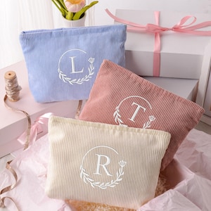 Op de afbeelding: Drie rechthoekige etuis van corduroy in lichtblauw, oudroze en crème. Elk etui is voorzien van een wit geborduurd cirkelvormig kransontwerp met een centrale letter: L op de blauwe, T op de roze en R op de crème etui. De etuis zijn samen gerangschikt.