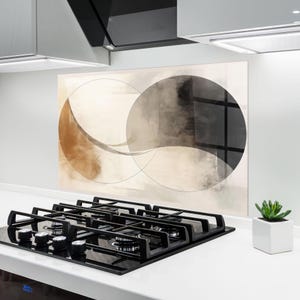 May include: Abstract art panel featuring overlapping circles in shades of brown, gray, and beige. The artwork is mounted on a white wall above a black stovetop with silver knobs. A small potted plant sits on the countertop.