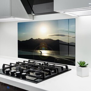 Op de afbeelding: Glazen keukenachterwand met een zonsondergang boven water en bergen, met een silhouet op het strand. De scène wordt omlijst door een wit aanrechtblad en een zwart gaskookplaat.