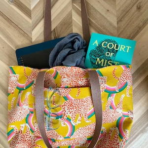 May include: Yellow tote bag with a leopard and rainbow pattern. The bag has brown straps and a pocket. Inside the bag are a book titled "A Court of Mist", a notebook, and a scarf. A pair of glasses rests on the pocket.