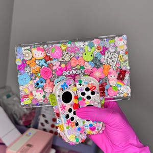 May include: A clear phone case and a controller case, both adorned with colorful charms, rhinestones, and the name "CHLOE". The cases feature various shapes, including flowers, bows, and cartoon characters, creating a playful and decorative design.