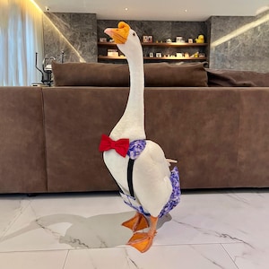 Puede incluir: Un ganso blanco con una pajarita roja, una prenda con estampado floral azul y morado y tirantes negros. El ganso tiene patas naranjas y un pico amarillo y naranja. El fondo incluye un sofá marrón y una estantería con objetos decorativos.