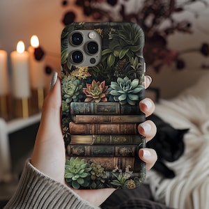 Op de afbeelding: Een telefoonhoesje met een ontwerp van een stapel vintage boeken met groene vetplanten die erop groeien. De hoes heeft een camera-uitsparing en een zwarte achtergrond.