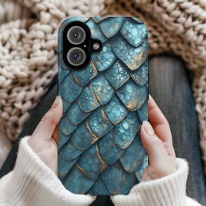 Puede incluir: Una funda de teléfono de color verde azulado y dorado con un diseño de escamas de dragón. La funda presenta un patrón texturizado con escamas superpuestas en tonos verde azulado y dorado. La funda del teléfono tiene un recorte para cámara de doble lente.
