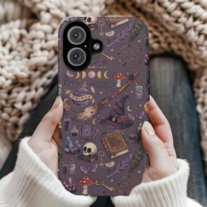 Peut inclure: Coque de téléphone avec un fond violet orné d'un motif de sorcière. Le motif comprend un crâne, un chapeau de sorcière, un balai, un livre de sorts, des clés, des champignons et des fioles de potion. L'étui a une découpe pour un appareil photo à double objectif.