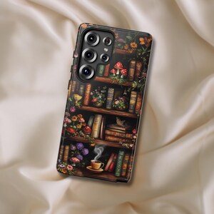 May include: A phone case with a whimsical design featuring a bookshelf filled with books, flowers, and mushrooms. A steaming cup of tea sits on the bottom shelf.