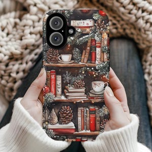 Peut inclure: Une coque de téléphone avec un motif hivernal représentant une bibliothèque remplie de livres, une tasse de café et des pommes de pin. La coque a un fond sombre avec un schéma de couleurs rouge, blanc et vert.