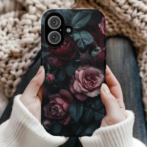 Puede incluir: Una funda de teléfono negra con un diseño floral con rosas de color rojo oscuro, rosa y verde. La funda tiene un recorte para el objetivo de la cámara.