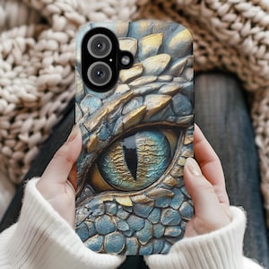 Könnte beinhalten: Detaillierte Handyhülle mit einem Drachenaugen-Design. Die Hülle hat ein strukturiertes, schuppiges Aussehen in Blau-, Gold- und Brauntönen. Das Auge ist ein auffälliger Blickfang mit einer blauen Iris und einer schwarzen Pupille. Die Hülle wird von einer Person in den Händen gehalten.