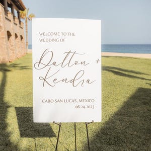 Może przedstawiać: Biały znak powitalny wesela na czarnym sztaludze. Znak głosi "Welcome to the Wedding of Dalton + Kendra" i "Cabo San Lucas, Mexico 06.24.2023". Znak znajduje się na trawniku, z plażą i morzem w tle.