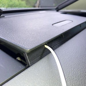 Op de afbeelding: Een close-up van een wit snoer dat door een zwart plastic compartiment in een autodashboard loopt.