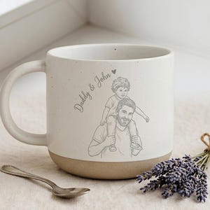 May include: Cream-colored ceramic mug with a tan base and a large handle. The mug features a sketch of a father carrying a child on his shoulders, with the words "Daddy & John" and a heart. A spoon and lavender sprigs are next to the mug.