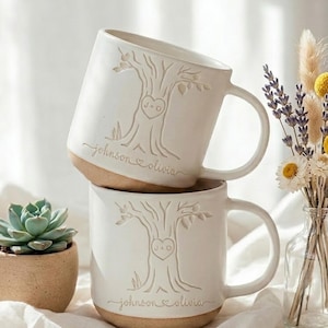 May include: Two stacked white ceramic mugs with a natural clay base, each featuring an embossed tree design with a heart containing the letters J+O and the text johnson + olivia. A small succulent in a tan pot and a glass vase with dried flowers.