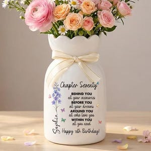 May include: A white ceramic vase with a cream-colored bow, filled with a bouquet of pink and peach roses, daisies, and other flowers. The vase has text that reads "Chapter Seventy" and "Happy 70th Birthday!"
