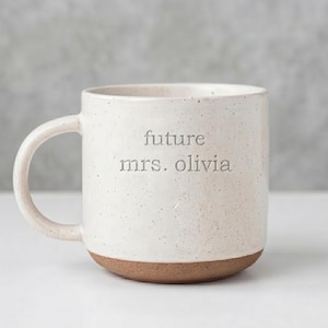 May include: A speckled off-white ceramic mug with a rounded handle and a natural brown clay base. The front of the mug features the debossed text "future mrs. olivia" in a simple, lowercase sans-serif font centred on the body of the vessel.