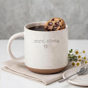 May include: A speckled off-white ceramic mug with a brown base, featuring the engraved text "mrs. olivia" and a small heart symbol. The mug is filled with a dark liquid and a chocolate chip cookie, with steam rising from the top of the mug.