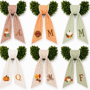 May include: Six decorative wall hangings featuring green boxwood foliage topped with fabric ribbons. The ribbons display various patterns including gingham and stripes, accented with embroidered seasonal motifs like pumpkins, turkeys, and letters.