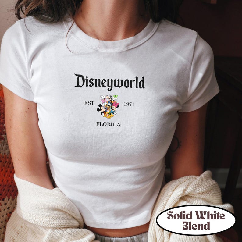 May include: White t-shirt with the text "Disneyworld" in a classic font, along with "EST 1971 FLORIDA" and a graphic of Disney characters. The shirt has a crew neck and short sleeves.