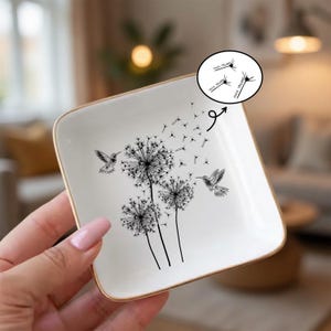 May include: Square white ceramic trinket dish with a gold rim. The dish features a black and white dandelion design with hummingbirds. The text "FUCK TRUMP" is in a speech bubble.