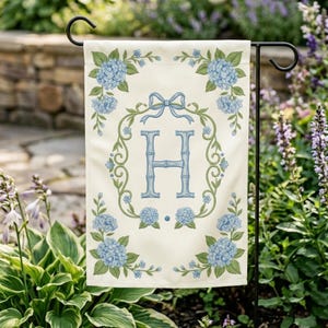 May include: A cream-colored garden flag featuring a blue floral design and the letter "H". The flag has a wreath of blue hydrangeas and green vines around the letter. A blue bow is at the top of the wreath. The flag is hanging on a black metal stand.