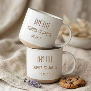 May include: Two off-white ceramic mugs stacked vertically, featuring a speckled texture and a natural clay base. Each mug is engraved with tally marks, the names Sophia and Jason separated by a heart symbol, and the date 09.06.17 in brown text.