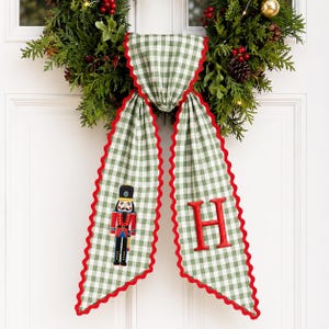 May include: A green and white gingham fabric door hanger featuring a red scalloped trim. One side displays an embroidered nutcracker figure in red, blue, and gold, while the other side features a large, embroidered red letter H in a serif font.