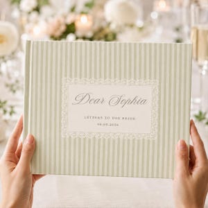 May include: A rectangular hardcover book with a light sage green and white vertical striped cover. The center features a white rectangular label with a decorative lace border, displaying the text Dear Sophia, LETTERS TO THE BRIDE, and 06.08.2026.