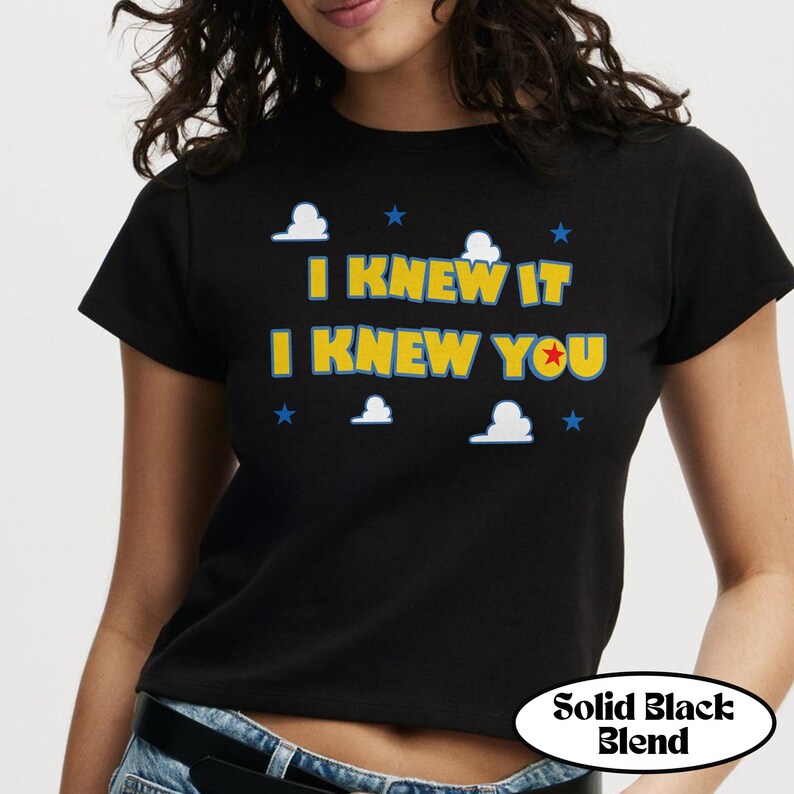 Puede incluir: Una camiseta negra de manga corta con el texto amarillo I KNEW IT I KNEW YOU y una estrella roja dentro de la letra O. El dise&ntilde;o incluye formas de nubes blancas y peque&ntilde;as estrellas azules dispersas alrededor del texto en la parte frontal.