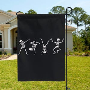 May include: Black garden flag featuring four white skeleton figures in various dance poses. The skeletons have text on their heads. The flag is hanging on a black metal pole in a grassy yard.