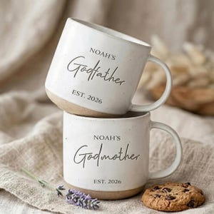 May include: Two off-white ceramic mugs with speckled textures and a natural clay base. The top mug features the text "NOAH'S Godfather EST. 2026" and the bottom mug features the text "NOAH'S Godmother EST. 2026" in dark, elegant lettering.