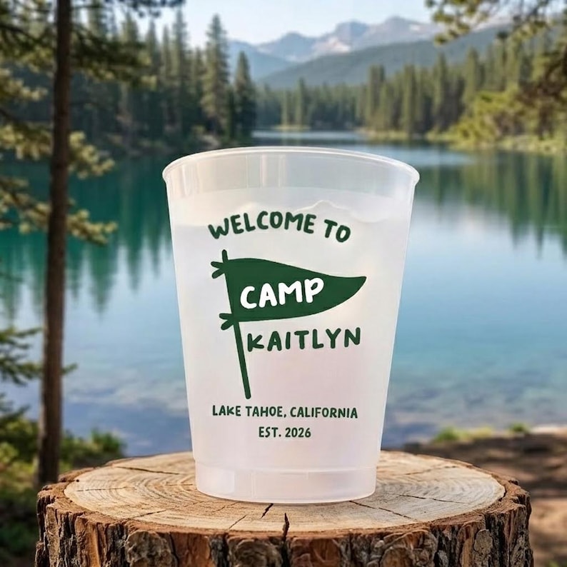 May include: A frosted plastic cup featuring dark green text that reads WELCOME TO, CAMP, KAITLYN, LAKE TAHOE, CALIFORNIA, EST. 2026, along with a graphic of a pennant flag. The cup is positioned on a textured wooden surface.