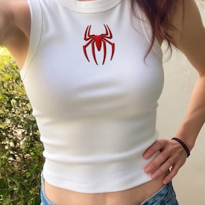 May include: White ribbed tank top with a red spider emblem on the chest. The sleeveless top is made of a soft material and is paired with blue denim jeans. The spider emblem is a detailed, stylized design.