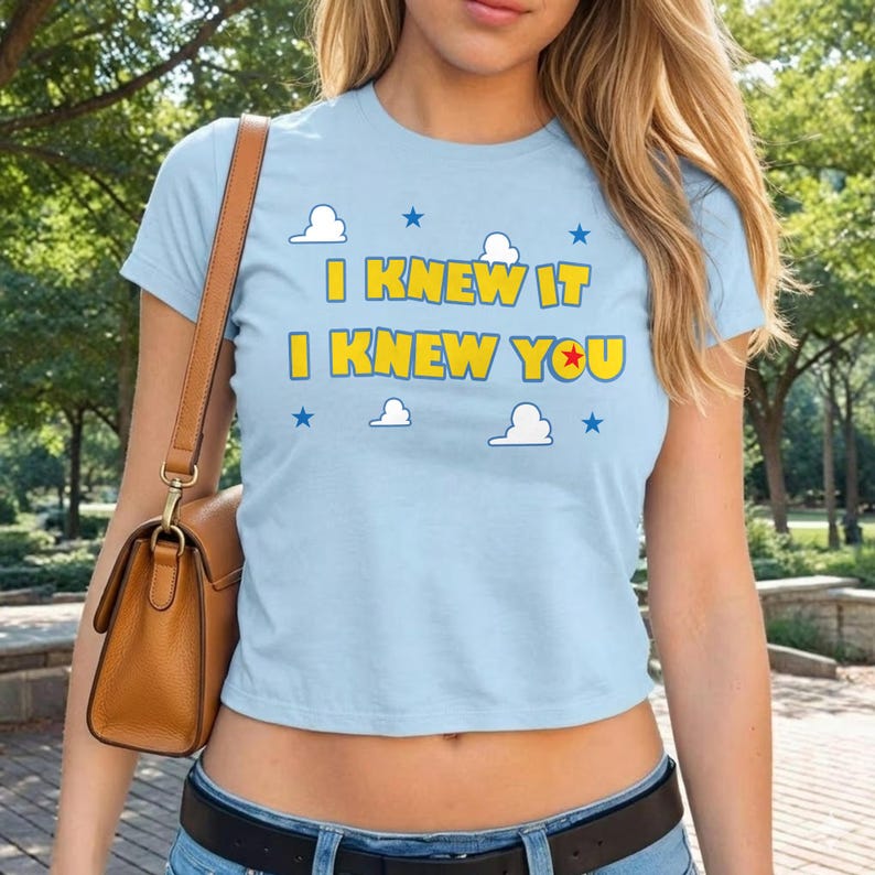 Puede incluir: Una camiseta de manga corta de color azul claro con el texto en amarillo I KNEW IT I KNEW YOU, donde una estrella roja sustituye a la letra O. El dise&ntilde;o incluye peque&ntilde;as nubes blancas y estrellas azules dispersas alrededor del texto.