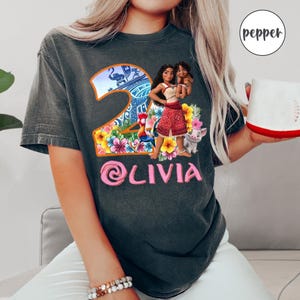 Peut inclure: Un t-shirt gris fonc&eacute; avec un motif color&eacute; de Moana et Pua du film Disney Moana. Le motif comprend un num&eacute;ro 2 avec des fleurs tropicales et un coq. Le texte "@livia" est imprim&eacute; en rose sous le motif.