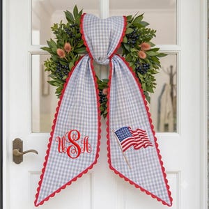 May include: A green leaf wreath with dark berries and peach-colored thistle accents, featuring a long light blue and white gingham fabric bow with red scalloped edges. The left tail displays red monogrammed letters USA, and the right tail shows an embroidered American flag.