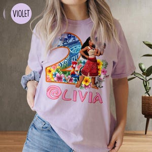 Peut inclure: T-shirt violet avec un motif de Moana de Moana de Disney, un coq et un cochon. Le motif est un num&eacute;ro 2 avec des fleurs et un design tropical. Le texte "Olivia" est sous le motif.