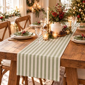 May include: A rectangular table runner featuring a pattern of vertical sage green and off-white stripes. The fabric runner is laid flat across the center of a wooden dining table, serving as a base for place settings, glassware, and a centerpiece.