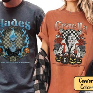 May include: Two t-shirts, one grey and one orange, with graphic designs. The grey shirt features "Hades" with a character illustration and text. The orange shirt displays "Cruella" with a character illustration, roses, and pumpkins.