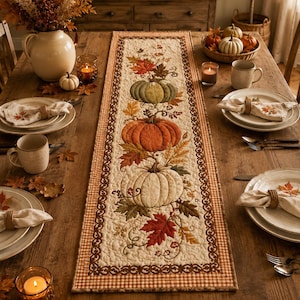 May include: A rectangular quilted table runner featuring a central design of three stacked pumpkins in green, orange, and white, surrounded by embroidered autumn leaves and vines, finished with a brown and white checkered border on a wooden table.