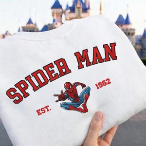 May include: White sweatshirt featuring the words "SPIDER MAN" in red block letters with a black outline. Below the text is a graphic of Spider-Man in his classic red and blue suit, with the year "1962" and the word "EST." in red.