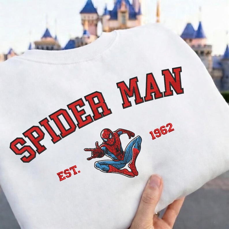 May include: White sweatshirt featuring the words "SPIDER MAN" in red block letters with a black outline. Below the text is a graphic of Spider-Man in his classic red and blue suit, with the year "1962" and the word "EST." in red.